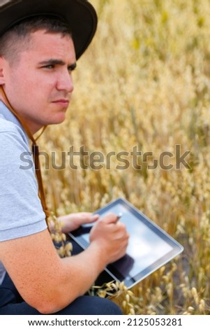 Young farmer. Portrait of farmer seating in gold wheat field and scrolling on tablet. Young man wearing cowboy hat in field examining wheat crop. Oats grain industry. Blurred. Oats plant. Copy space.