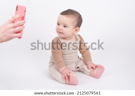 Baby wearing beige overalls sitting on white background looking at hand holding a telephone and watching cartoons. 
