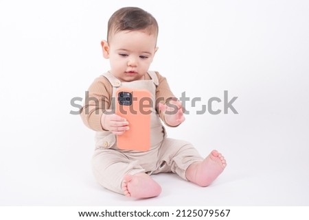 Baby wearing beige overalls sitting on white background holding a telephone and watching cartoons. 