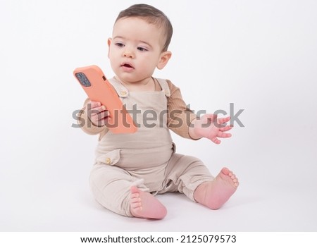 Baby wearing beige overalls sitting on white background holding a telephone and watching cartoons or movies. 