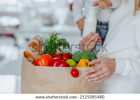 Young mother in white hoodie holding a baby girl and posing with paper package of fresh vegetables, milk and bread. Focus is at the package. Delivery food concept. Royalty-Free Stock Photo #2125085480