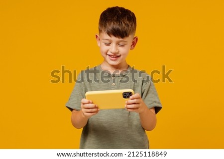 Little small boy 6-7 years old in green t-shirt using play racing app on mobile cell phone gadget smartphone for pc video games isolated on plain yellow background. Mother's Day love family concept