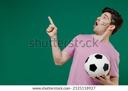 Young fun man fan wears basic pink t-shirt cheer up support football sport team hold in hand soccer ball watch tv live stream scream point on workspace isolated on dark green color background studio