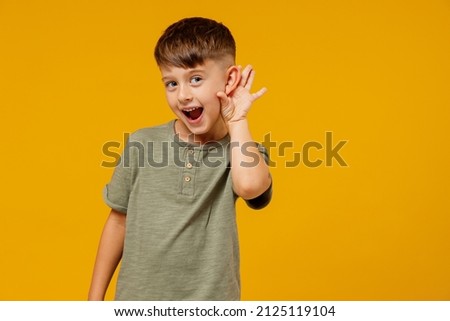 Little small smiling happy boy 6-7 years old in green t-shirt try to hear you overhear listen intently isolated on plain yellow background studio portrait. Mother's Day love family lifestyle concept Royalty-Free Stock Photo #2125119104