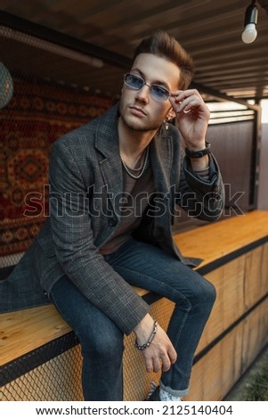 Fashionable vogue hipster man in stylish casual coat and jeans sits and wears a sunglasses in the city Royalty-Free Stock Photo #2125140404