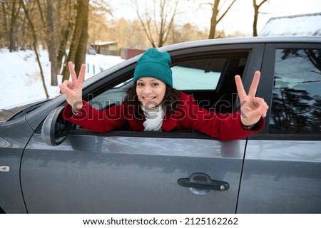 Cheerful gorgeous Caucasian woman driver traveler travelling by car on a snowy woodland, leaning on the driver door with opened windows and showing peace signs, cutely smiles looking at camera.