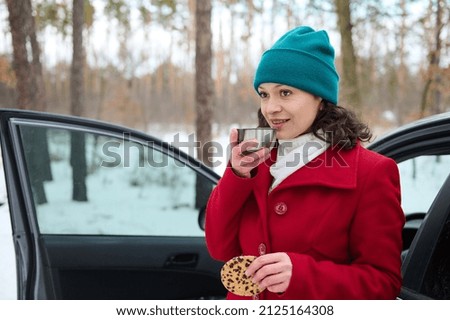 Middle aged African American woman taking her lunch outdoor, standing near a driver door of her automobile, resting from the road in the snow covered nature while travelling by car at winter holiday