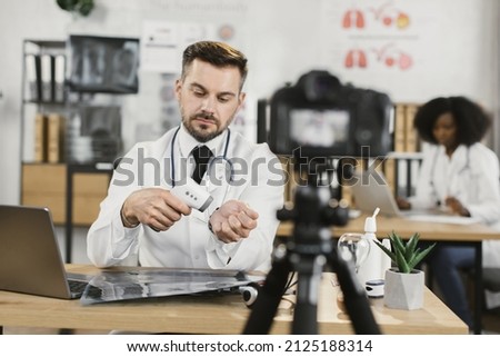 Positive caucasian doctor in lab coat holding white sterile gloves in hands and talking on camera while recording video. Concept of medicine and social media.