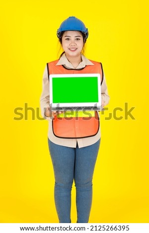 Portrait studio shot of Asian female professional engineering foreman manager in hard helmet and reflect safety vest hold blank green screen laptop computer for text copy space on yellow background.