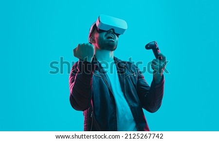 Excited male gamer in VR headset and with joystick celebrating victory in videogame in cyberspace on blue background in studio with pink neon light
