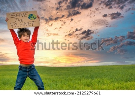 6 Year Old Boy Holding A Cardboard Sign That Says SAVE THE PLANET. He Is Jumping On A Green Meadow With A Dramatic Sky And A Nice Horizon. He Wears A Red Shirt And Jeans. Image with copy space