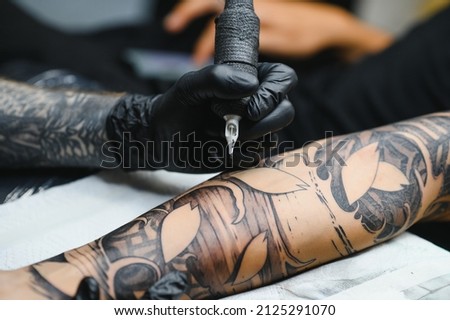 Cropped close up of a bearded tattoo artist working at his studio tattooing sleeve on the arm of his male client. Man getting tattooed by professional tattooist Royalty-Free Stock Photo #2125291070