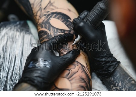 Cropped close up of a bearded tattoo artist working at his studio tattooing sleeve on the arm of his male client. Man getting tattooed by professional tattooist Royalty-Free Stock Photo #2125291073