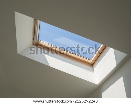 Dormer wooden window in the white sloping ceiling overlooking the blue sky. Sunlight enters the room through a closed window Royalty-Free Stock Photo #2125292243
