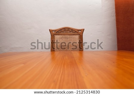 An empty wooden chair at an empty wooden table, an uneven wall and a piece of curtain in the background, perspective.