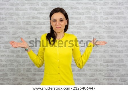 Portrait of a confused casual girl shrugging shoulders isolated over grey brick wall background. woman with puzzled expression, spreads hands with confused look, frowns face. Doubt concept