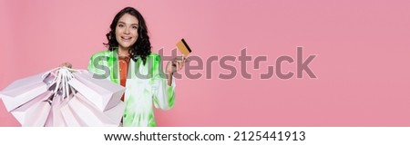 cheerful young woman in tie dye blazer holding credit card and shopping bags isolated on pink, banner