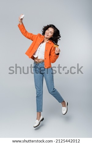 full length of young woman in orange jacket holding credit card with cashback and taking selfie while levitating on grey