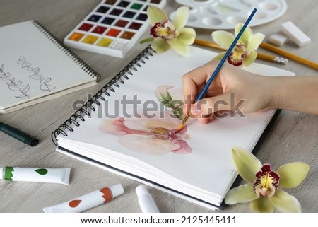 Woman drawing beautiful orchid flowers in sketchbook at wooden table, closeup