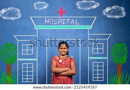 Happy smiling Indian girl kid with stethoscope standing infront of Hospital painting - concept of poor child dreaming about doctor as future career and education. Royalty-Free Stock Photo #2125459187