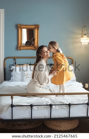 Little girl kissing her loving mom, sitting on bed at cozy bedroom with modern retro interior. The concept of a happy relationship between mother and daughter. Happy mothers day. High quality photo