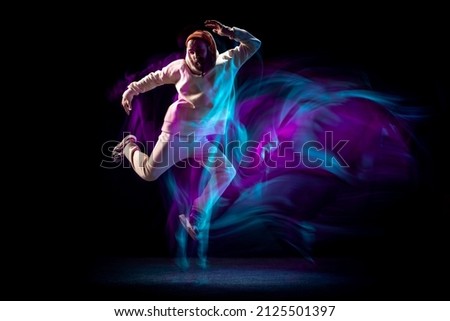 Jumping. Energy young sportive man dancing hip-hop or breakdance in white outfit on dark background in mixed blue neon light. Concept of sport, art, action, moves, youth culture. Banner for ad