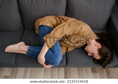 Top view of tired unhappy young female lying on couch curled up suffers from menstrual cramps. Unplanned pregnancy and abortion, try to sleep hiding from life troubles, break up and divorce concept Royalty-Free Stock Photo #2125514618