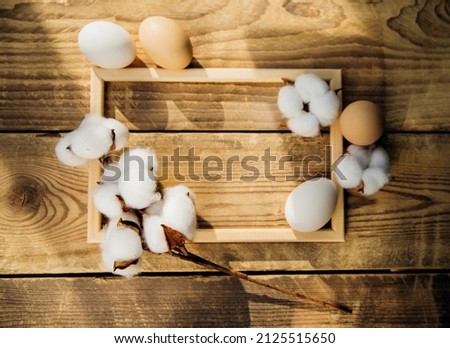 Eggs and a branch with white cotton flowers with sunny shadows on a wooden background . Twigs are intertwined with a wooden frame. A holiday card. The Concept of the Easter Holiday.