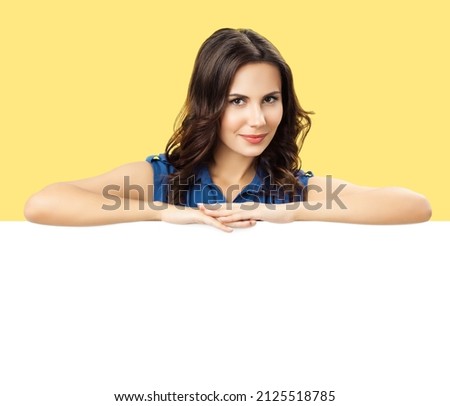 Portrait image of smiling beautiful brunette woman in blue dress cloth, standing behind, peeping from blank banner mock up mockup signboard with copy space, over yellow color background. Royalty-Free Stock Photo #2125518785