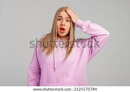 Yikes, oops. Young blonde woman wears violet hoody, stands against grey background surprised with hand on head for mistake, remember error. Forgot, bad memory concept Royalty-Free Stock Photo #2125570784