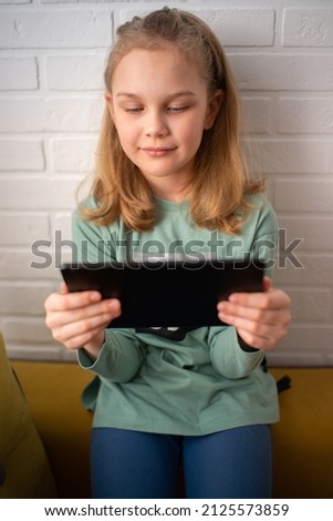 Smiling little girl is sitting on the armrest of the sofa and enjoying playing an online game on a digital tablet computer. Addicted to technology, web surfing information.