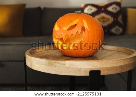 Carved Halloween pumpkin, jack lantern (Jack-o'-lantern). Spooky laughing, scary head on a small table in a home living room, with a couch in the background.