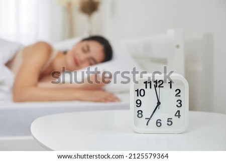 Alarm clock that is going to ring at seven in morning on background of woman sleeping in bed. Close up of alarm clock standing on table next to young woman sleeping sweetly in her bed. Selective focus Royalty-Free Stock Photo #2125579364