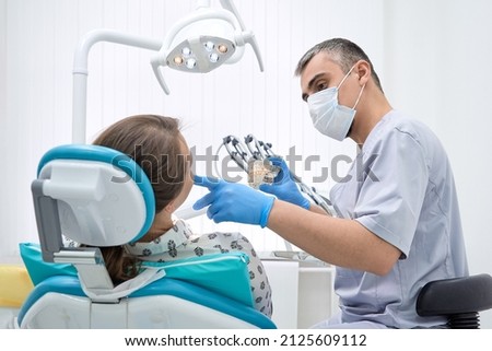An orthodontist conducts a consultation on dental prosthetics. Doctor dentist shows an artificial plastic jaw with dental implants. Background of modern clinic Royalty-Free Stock Photo #2125609112
