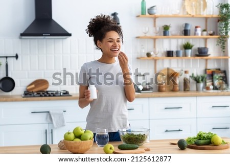 Happy african american woman standing at the cuisine table in the home kitchen drinking dietary supplements, looking away and smiling friendly, healthy lifestyle concept Royalty-Free Stock Photo #2125612877