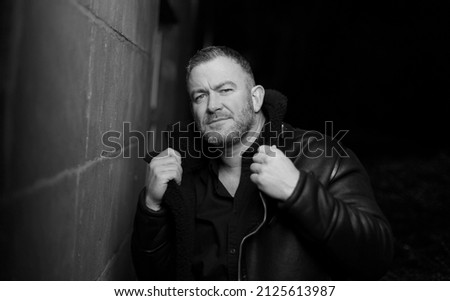 Caucasian male standing next to wall and posing with his leather coat.