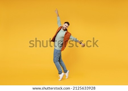 Full length young smiling happy confident smiling cheerful fun caucasian man 20s wearing orange vest mint sweatshirt stand on toes leaning back dancing isolated on yellow background studio portrait.