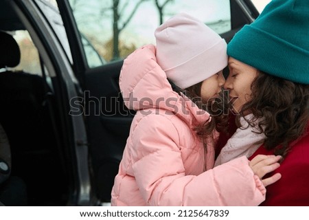 Close-up portrait a beautiful woman- delightful mom and daughter hugging each other standing close to each other face to face near the family automobile during winter trip by car