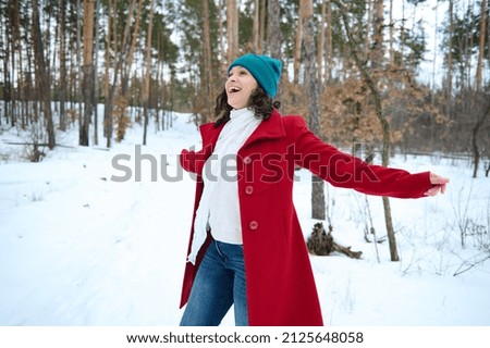 Attractive middle aged woman in bright colorful warm clothes and white woolen scarf whirling around herself, smiles, rejoices, enjoying her weekend in the snow covered forest on a sunny snowy day Royalty-Free Stock Photo #2125648058