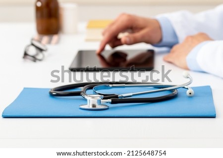 Closeup shot of a stethoscope resting on a tablet with an unrecognizable doctor working in the background with laptop