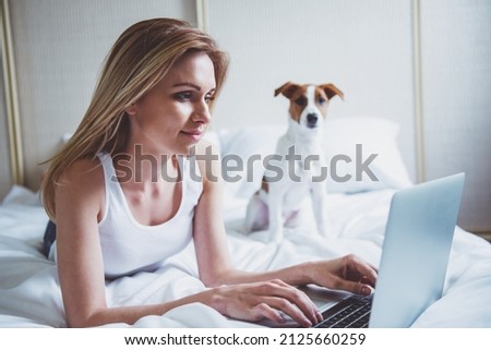 Beautiful blonde girl in casual clothes is using a laptop and smiling while lying with her cute dog on bed. Pet is looking at camera