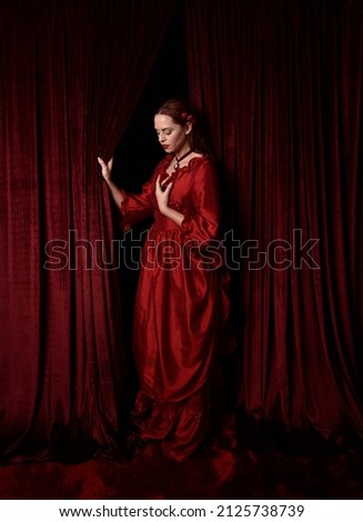  portrait of pretty female model with red hair wearing glamorous historical victorian red ballgown.  Posing with a moody dark background.
