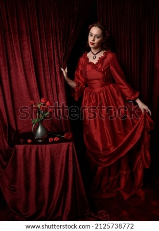  portrait of pretty female model with red hair wearing glamorous historical victorian red ballgown.  Posing with a moody dark background.