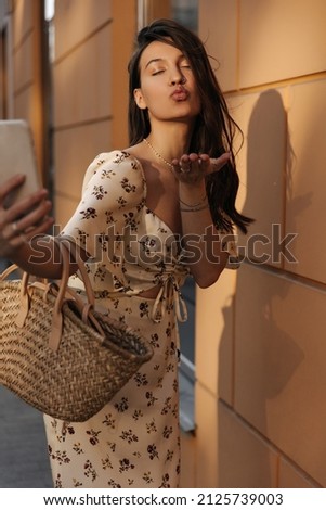 Pretty young woman sends air kiss while standing outdoors against background of wall. Brown-haired with her eyes closed takes pictures on phone, dressed in light white outfit. Cell phone use concept