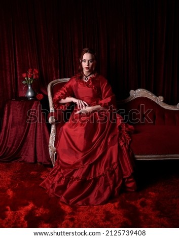  portrait of pretty female model with red hair wearing glamorous historical victorian red ballgown.  Posing with a moody dark background, sitting on  ornate chair. Royalty-Free Stock Photo #2125739408