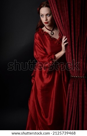  portrait of pretty female model with red hair wearing glamorous historical victorian red ballgown.  Posing with a moody dark background, sitting on  ornate chair. Royalty-Free Stock Photo #2125739468