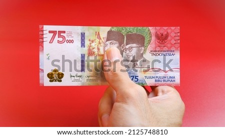Indonesian Rupiah the official currency of Indonesia. A man's hand is making a payment. Male hand showing Indonesian Rupiah note. Business and finance concept. Red Background. Uang 75000 Rupiah.