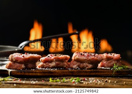 Raw steaks meat beef with seasoning on wooden chopping board on a wooden table prepared for cooking with flames in the background.