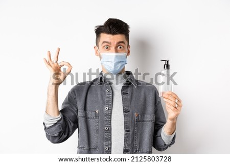 Healthy people and covid-19 concept. Excited man in medical mask holding bottle of good hand sanitizer, show okay sign, recommend antiseptic, standing against white background