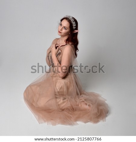 Full length portrait of pretty female model with red hair wearing glamorous fantasy tulle gown, crown and shroud veil.  Posing in a seated kneeling pose on a studio background